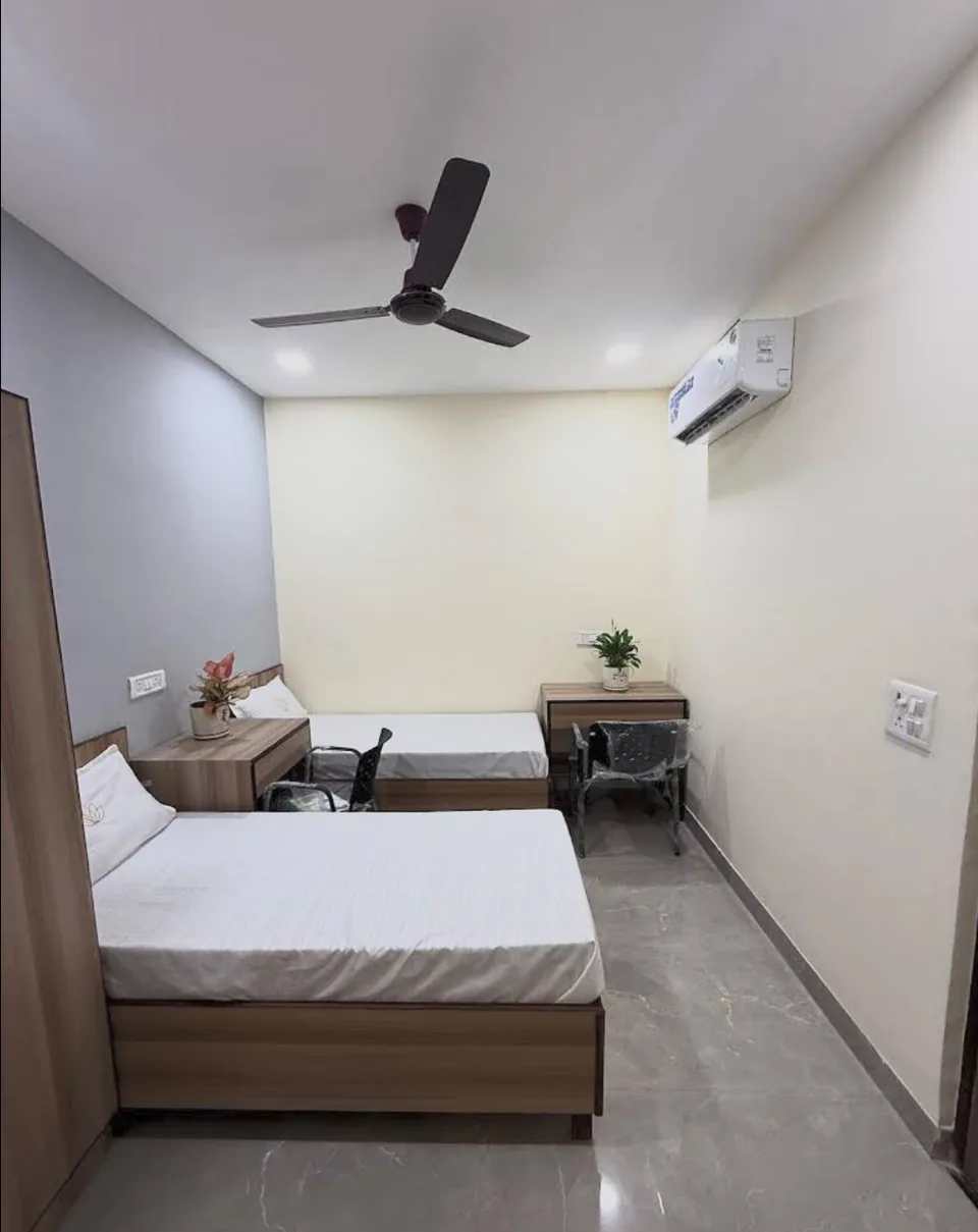 Twin-sharing room with two beds, study desks, ceiling fan and wall-mounted AC.